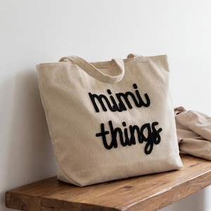 May include: A beige corduroy tote bag with the words "mimi things" in black script. The bag has a rectangular shape with a handle and is sitting on a wooden bench. A tan coat is draped over the bench.