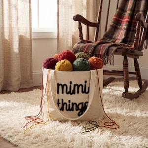 K&ouml;nnte beinhalten: Eine cremefarbene Tragetasche mit den schwarzen Worten "mimi things" ist mit bunten Wollkn&auml;ueln gef&uuml;llt. Die Tasche steht auf einem flauschigen wei&szlig;en Teppich, wobei Garnstr&auml;nge herausragen. Ein Schaukelstuhl aus Holz und eine karierte Decke befinden sich im Hintergrund.