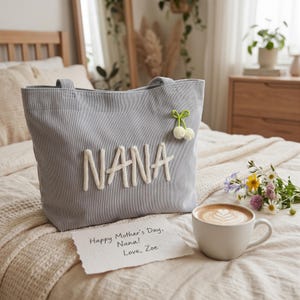 K&ouml;nnte beinhalten: Eine graue Cord-Tragetasche mit dem Wort "NANA" in Wei&szlig; gestickt. Eine kleine, dekorative Kirsche ist an der Tasche befestigt. Eine handgeschriebene Notiz lautet "Happy Mother's Day, Nana! Love, Zoe." Eine Tasse Kaffee mit Latte Art steht daneben.