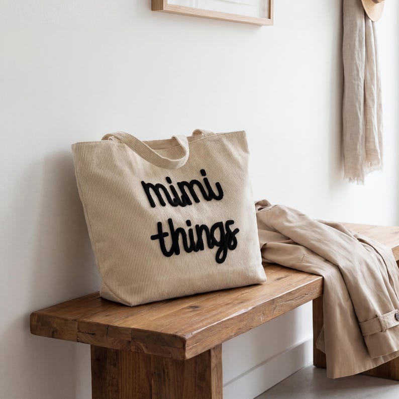 May include: A beige corduroy tote bag with the words "mimi things" in black script. The bag has a rectangular shape with a handle and is sitting on a wooden bench. A tan coat is draped over the bench.