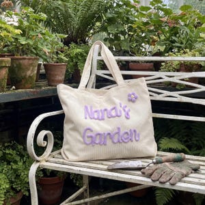K&ouml;nnte beinhalten: Cremefarbene Cord-Tragetasche mit dem gestickten Text "Nana's Garden" in Lavendel. Eine kleine Lavendelblume ist oben rechts aufgen&auml;ht. Die Tasche hat zwei Henkel und liegt auf einer wei&szlig;en Bank mit Gartenger&auml;ten.