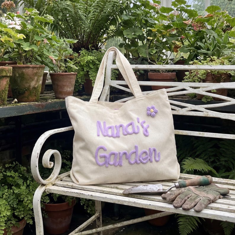 K&ouml;nnte beinhalten: Cremefarbene Cord-Tragetasche mit dem gestickten Text "Nana's Garden" in Lavendel. Eine kleine Lavendelblume ist oben rechts aufgen&auml;ht. Die Tasche hat zwei Henkel und liegt auf einer wei&szlig;en Bank mit Gartenger&auml;ten.