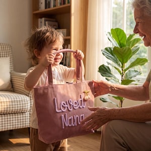 K&ouml;nnte beinhalten: Eine rosa Cord-Einkaufstasche mit dem gestickten Text "Loved Nana". Die Tasche hat zwei Griffe und eine kleine Kirschverzierung. Ein Kind h&auml;lt die Tasche.