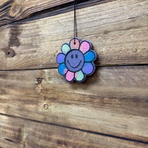 May include: A colorful, flower-shaped air freshener with a smiling face. The petals are various colors, including pink, blue, and purple, with glitter accents. It hangs from a black string against a wooden background.