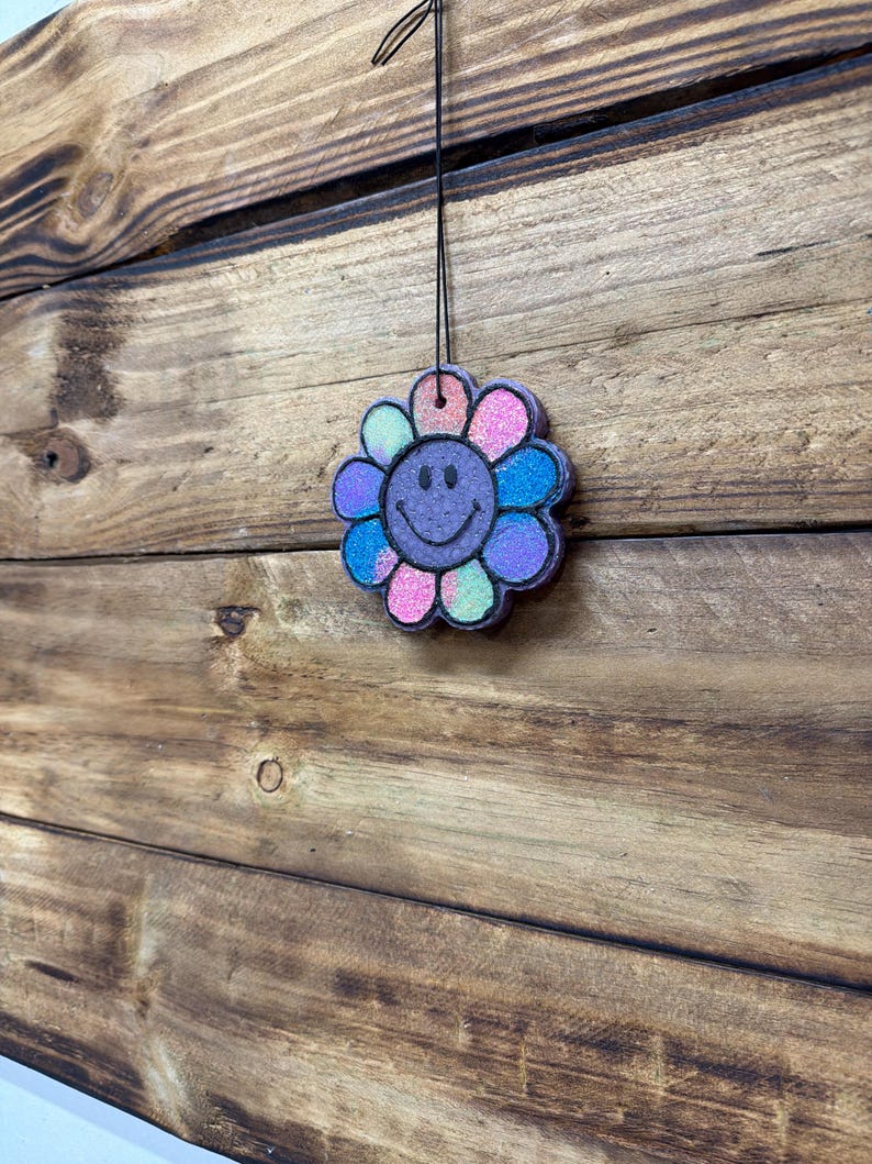 May include: A colorful, flower-shaped air freshener with a smiling face. The petals are various colors, including pink, blue, and purple, with glitter accents. It hangs from a black string against a wooden background.