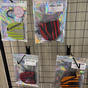 May include: Several air fresheners in holographic bags are displayed on a black grid. The air fresheners have various designs, including the word "Cummins" and a red and black striped design. Each bag has a label with the scent name.