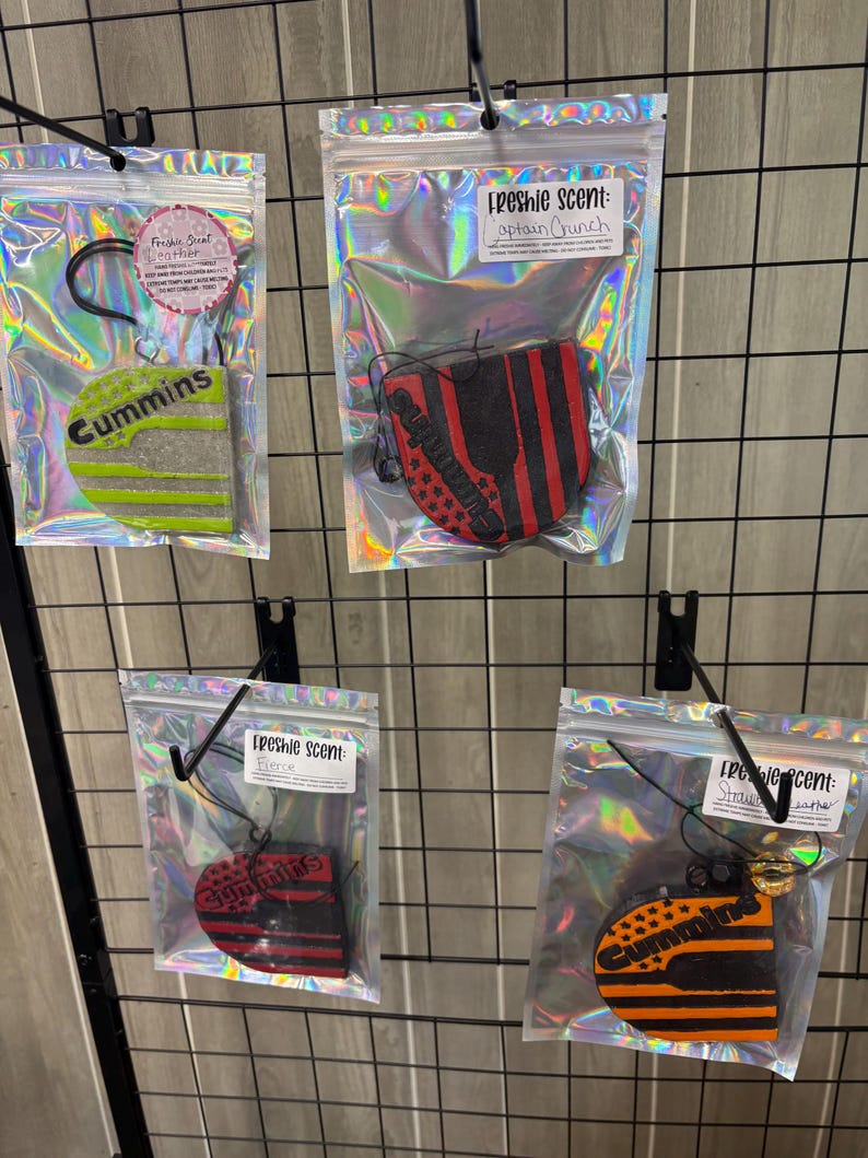 May include: Several air fresheners in holographic bags are displayed on a black grid. The air fresheners have various designs, including the word "Cummins" and a red and black striped design. Each bag has a label with the scent name.