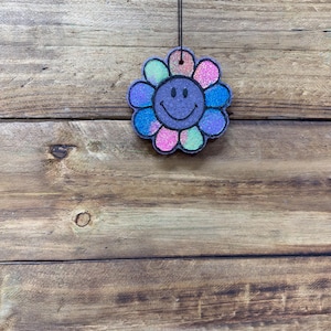May include: A colorful, glittery flower-shaped air freshener with a smiling face. The flower petals are a mix of pink, blue, green, and purple. It hangs from a black string against a wooden background.
