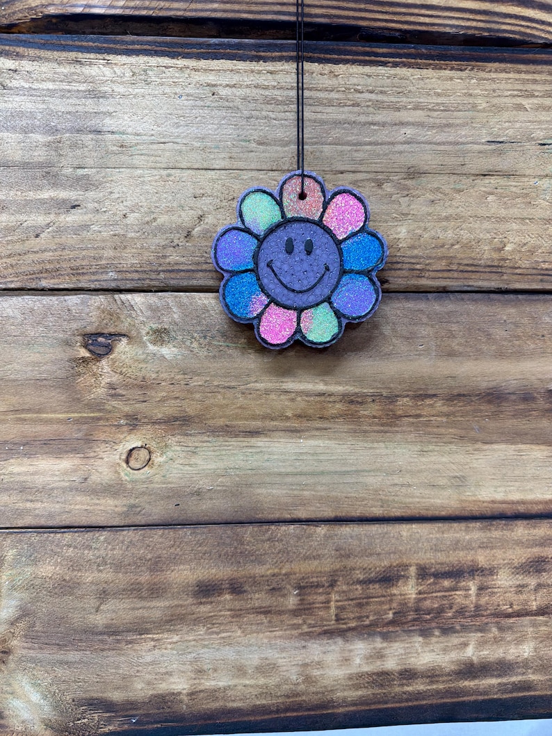 May include: A colorful, glittery flower-shaped air freshener with a smiling face. The flower petals are a mix of pink, blue, green, and purple. It hangs from a black string against a wooden background.
