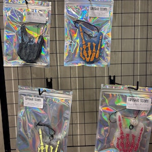 May include: Four air fresheners in holographic bags, each shaped like a hand making a rock-and-roll gesture. The fresheners are black, orange, yellow, and pink, with the text "Freshie scent" on each label.
