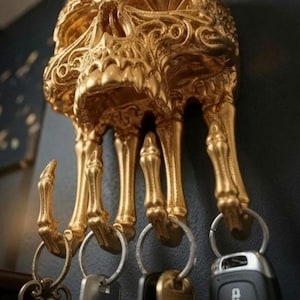 May include: A gold-coloured wall-mounted hook rack shaped like a human skull with skeletal fingers extending downwards to form four individual hooks. Each hook holds a metal key ring with various keys and a black electronic car key fob attached.