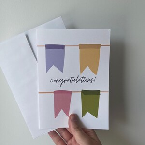 Puede incluir: Una tarjeta de felicitación blanca con la palabra "¡felicidades!" y cuatro banderines de colores. Las banderas son moradas, doradas, rosas y verdes. Un sobre blanco está al lado de la tarjeta.