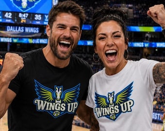 May include: A black t-shirt and a white t-shirt, each featuring a graphic of a winged horse head above a basketball and the text WINGS UP in blue, yellow, and white. The shirts are short-sleeved with a crew neckline and a screen-printed design.