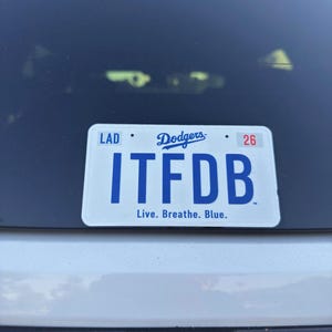 May include: A white rectangular license plate frame featuring the blue Dodgers logo at the top center, the text LAD on the left, the number 26 on the right, the large blue letters ITFDB in the center, and the text Live. Breathe. Blue. at the bottom.