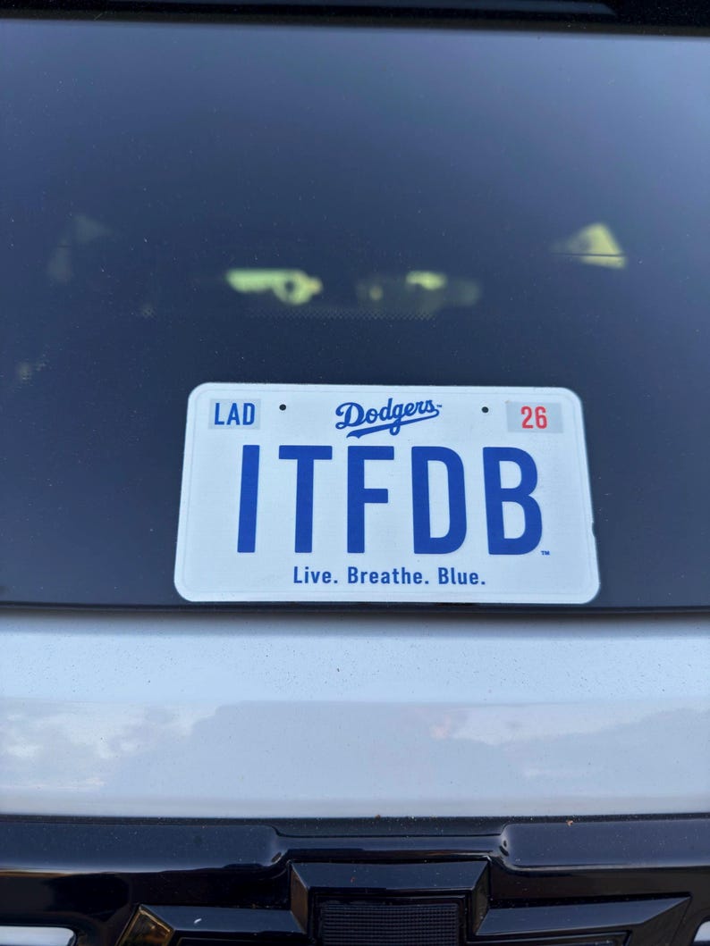 May include: A white rectangular license plate frame featuring the blue Dodgers logo at the top center, the text LAD on the left, the number 26 on the right, the large blue letters ITFDB in the center, and the text Live. Breathe. Blue. at the bottom.