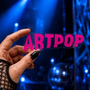 May include: A clear, die-cut sticker featuring the word ARTPOP printed in a bold, vibrant hot pink sans-serif font. The sticker has a transparent background and is held up to display the lettering clearly against a dark, out-of-focus blue background.