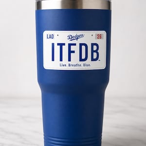 May include: A royal blue stainless steel tumbler with a clear lid and a white rectangular graphic resembling a license plate. The graphic features the text LAD, Dodgers, 26, ITFDB, and the phrase Live. Breathe. Blue. in blue and red lettering.