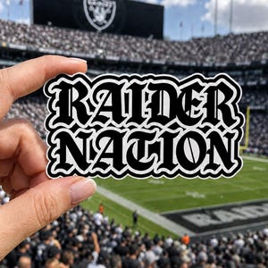 May include: A die-cut sticker featuring the words RAIDER NATION in a bold, black, gothic-style font with a thick white outline. The lettering is arranged in two stacked lines with a wavy, contoured border following the shape of the text.