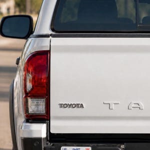 May include: The rear view of a white Toyota Tacoma pickup truck featuring a silver Toyota emblem and partial Tacoma lettering on the tailgate. A license plate is attached to the black bumper, displaying the text ITFDB, Dodgers, and Live. Breathe. Blue.