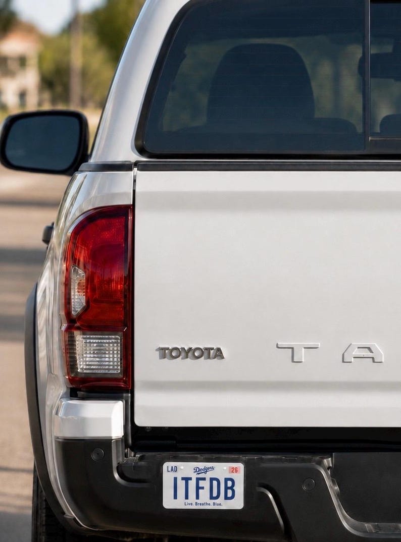 May include: The rear view of a white Toyota Tacoma pickup truck featuring a silver Toyota emblem and partial Tacoma lettering on the tailgate. A license plate is attached to the black bumper, displaying the text ITFDB, Dodgers, and Live. Breathe. Blue.
