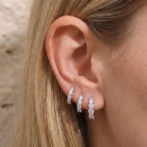 May include: Three silver-toned hoop earrings arranged in a row along the earlobe. Each hoop features a series of small, clear, round-cut stones set along the front-facing edge, creating a sparkling, continuous line of light against the metal.