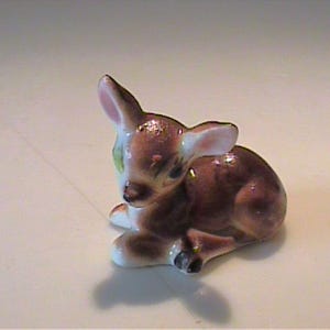 May include: A small ceramic figurine of a resting fawn with a glossy finish. The deer is colored in shades of brown and white, featuring large ears, dark eyes, and a small black nose. It is depicted in a lying position with its legs tucked underneath.