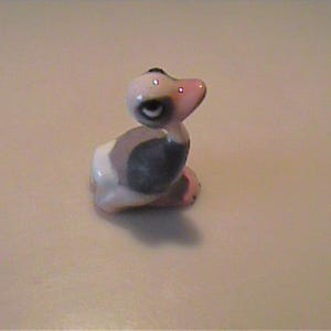 May include: A small ceramic figurine of a duckling with a white head, a dark grey body, and a light pink beak. The duckling is depicted in a sitting position with its head turned slightly to the side, featuring painted black eyes and subtle details.