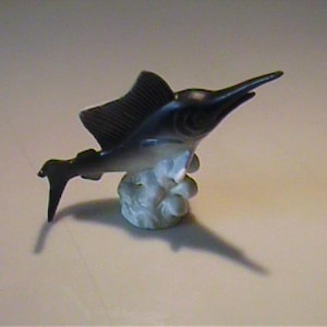 May include: A small, decorative figurine of a sailfish. The fish is primarily dark blue and white, with a prominent dorsal fin and a long, pointed bill. It is mounted on a white, cloud-like base.