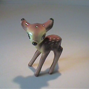 May include: A small ceramic figurine of a fawn with a glossy finish. The deer has a light tan face, dark brown ears with black tips, and a dark brown body featuring white spots along the back. It has large, dark eyes and a black nose.