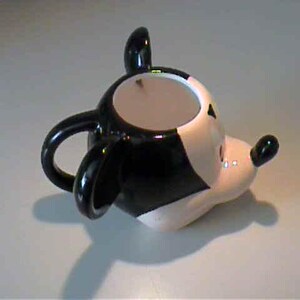 May include: A ceramic mug shaped like the head of a cartoon mouse. The mug features a glossy black and white color scheme, large rounded ears, a protruding snout with a small black nose, and a curved handle attached to the back of the head.
