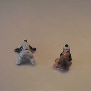 May include: Two miniature ceramic dog figurines. One figurine is white with black ears and a black tail, shown from the rear. The second figurine is brown, black, and white, depicted in a seated position facing away from the viewer.