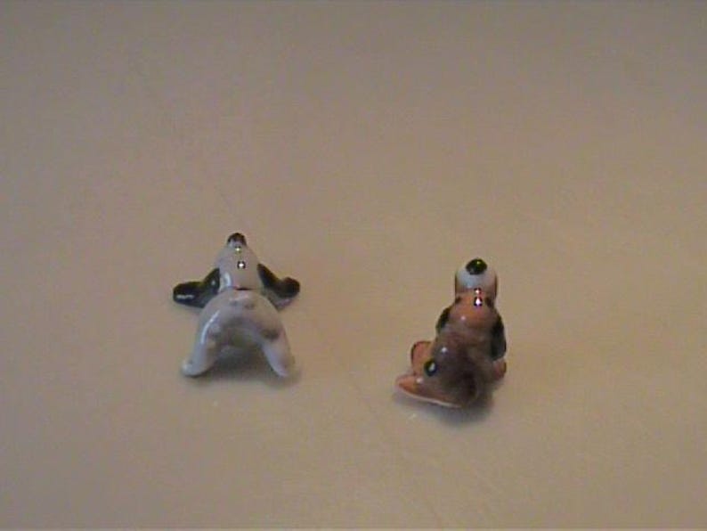 May include: Two miniature ceramic dog figurines. One figurine is white with black ears and a black tail, shown from the rear. The second figurine is brown, black, and white, depicted in a seated position facing away from the viewer.