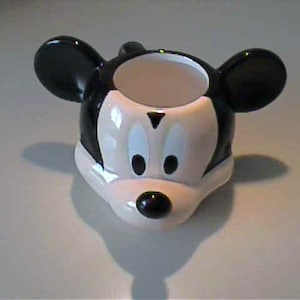 May include: A ceramic egg cup shaped like the head of Mickey Mouse, featuring a glossy black and white finish. The design includes two prominent rounded ears, a white face area with black oval eyes, and a small black nose at the center.