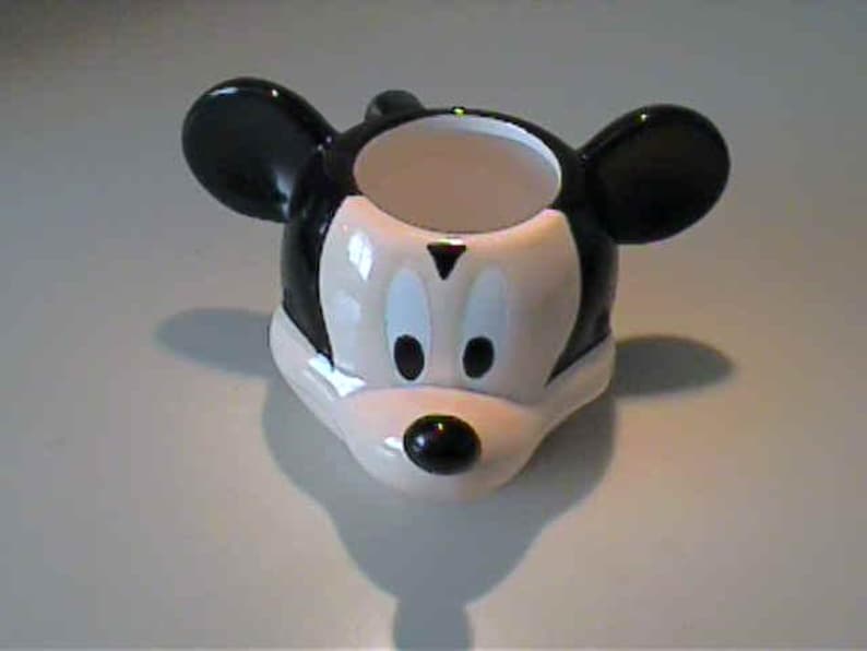 May include: A ceramic egg cup shaped like the head of Mickey Mouse, featuring a glossy black and white finish. The design includes two prominent rounded ears, a white face area with black oval eyes, and a small black nose at the center.
