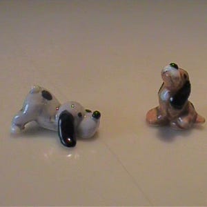 May include: Two small ceramic dog figurines. One figurine is light gray with black spots and long black ears, depicted in a lying down position. The second figurine is brown and white with black ears, shown in a sitting position looking upward.