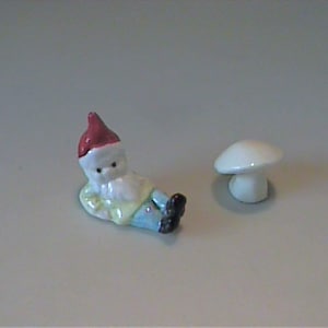 May include: A small ceramic figurine of a gnome wearing a red hat, a light yellow shirt, and blue pants, depicted in a reclining position. Beside the gnome is a separate, small, plain white ceramic mushroom figurine with a rounded cap.