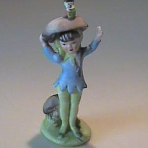 May include: A ceramic figurine of a fairy wearing a blue dress and yellow leggings, holding a mushroom over its head. A smaller figure sits atop the mushroom. The figurine stands on a green base with a mushroom and grass.