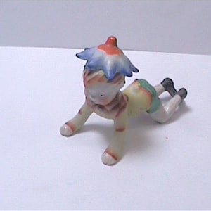 Könnte beinhalten: Keramikfigur eines krabbelnden Kindes mit einem blütenförmigen Hut. Die Figur hat einen rot-blauen Hut, ein gestreiftes Hemd und grüne Shorts. Das Kind ist auf Händen und Knien, mit schwarzen Schuhen.