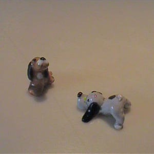 May include: Two small ceramic dog figurines. One figurine is brown and black, depicted in a sitting position. The second figurine is white with black spots and black ears, shown in a lying down position with its head resting on the ground.