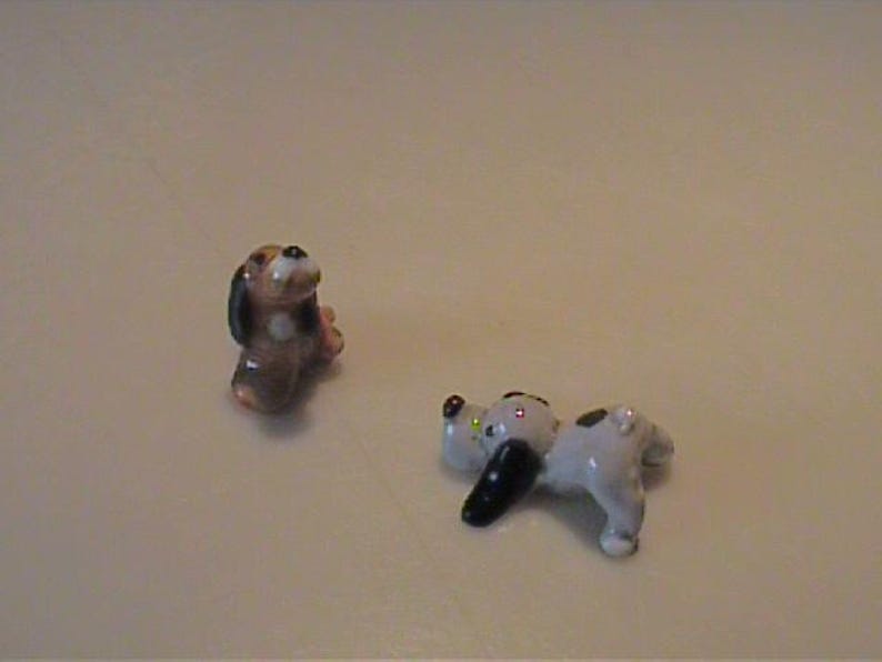 May include: Two small ceramic dog figurines. One figurine is brown and black, depicted in a sitting position. The second figurine is white with black spots and black ears, shown in a lying down position with its head resting on the ground.