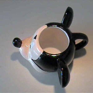 May include: A ceramic mug shaped like a cartoon mouse head, featuring a glossy black and white finish. The mug has a rounded body, a protruding snout with a black nose, two large black ears, and a small handle on the side for holding.