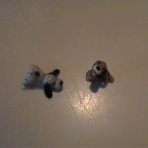 May include: Two miniature ceramic dog figurines. One figurine is white with black spots and black ears, depicted in a lying position. The second figurine is brown and white with a dark face, shown in a seated position with its head tilted upward.