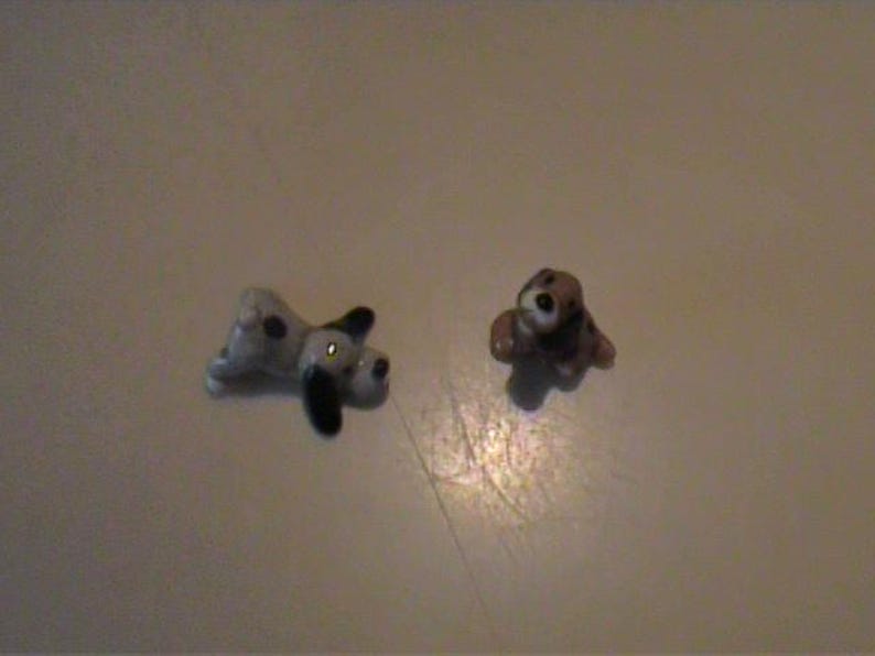 May include: Two miniature ceramic dog figurines. One figurine is white with black spots and black ears, depicted in a lying position. The second figurine is brown and white with a dark face, shown in a seated position with its head tilted upward.