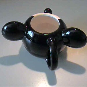 May include: A glossy black ceramic mug featuring a rounded body with three protruding, bulbous black handles extending from the sides. The interior of the mug is white, and the top rim is also white, contrasting with the dark exterior finish.