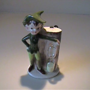 Może przedstawiać: Ceramiczna figurka chłopca w zielonym kapeluszu i stroju, stojącego obok pnia drzewa z klepsydrą. Figurka ma białą podstawę i jest pomalowana ze szczegółami.