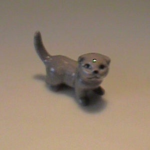 Pode incluir: Pequena figura de cerâmica cinzenta de uma lontra. A lontra é retratada numa pose de caminhada com a cauda curvada para cima. A figura tem um acabamento liso e detalhes faciais.