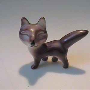 May include: A small, ceramic fox figurine in shades of purple and gray. The fox has its eyes closed, with a small black nose and a curved tail. The figurine is standing on a white surface.