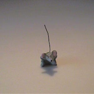 May include: A small gray mouse figurine with pink-tinted ears and a thin, dark, upright wire tail. The mouse has a pointed snout, visible eyes, and a compact body shape, presented as a singular, detailed miniature animal sculpture.