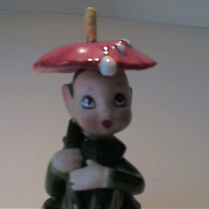 May include: A ceramic figurine of a small elf-like character wearing a wide, red, mushroom-shaped hat with white spots. The figure has a pale face with painted features, dark green clothing, and is holding a dark green umbrella-like accessory.