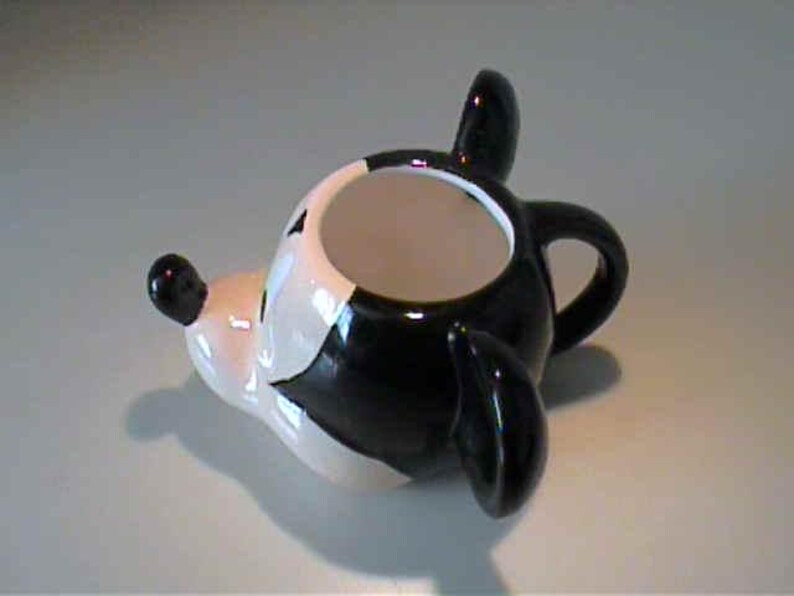 May include: A ceramic mug shaped like a cartoon mouse head, featuring a glossy black and white finish. The mug has a prominent snout with a black nose, two large rounded ears, and a small handle on the back. The interior of the mug is white.